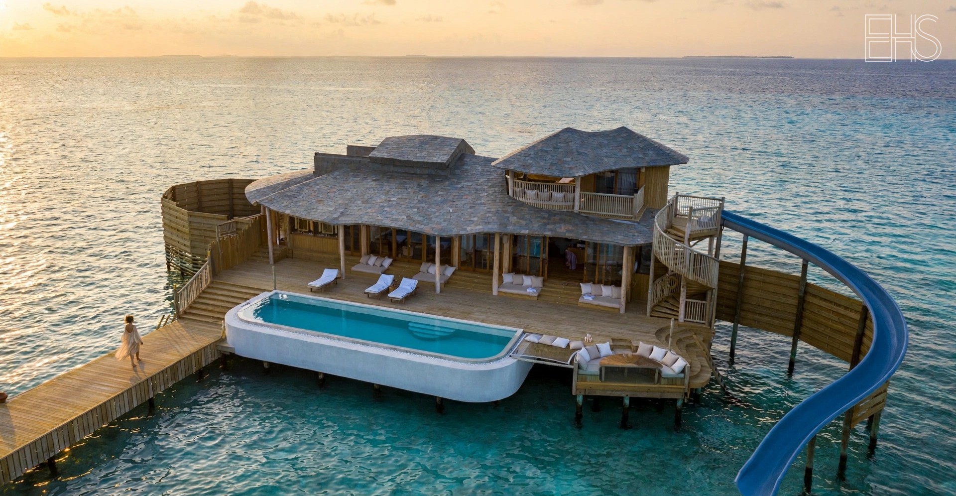 Soneva Fushi's overwater Water Retreat with water slide into the lagoon at golden hour, a woman walking the wooden jetty